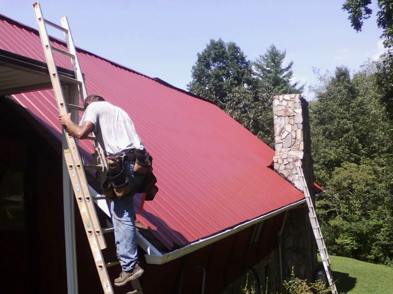 Roofer on a ladder beside a red metal roof and stone chimney — Soffit & Fascia Repair by Roofing Brothers in Leola